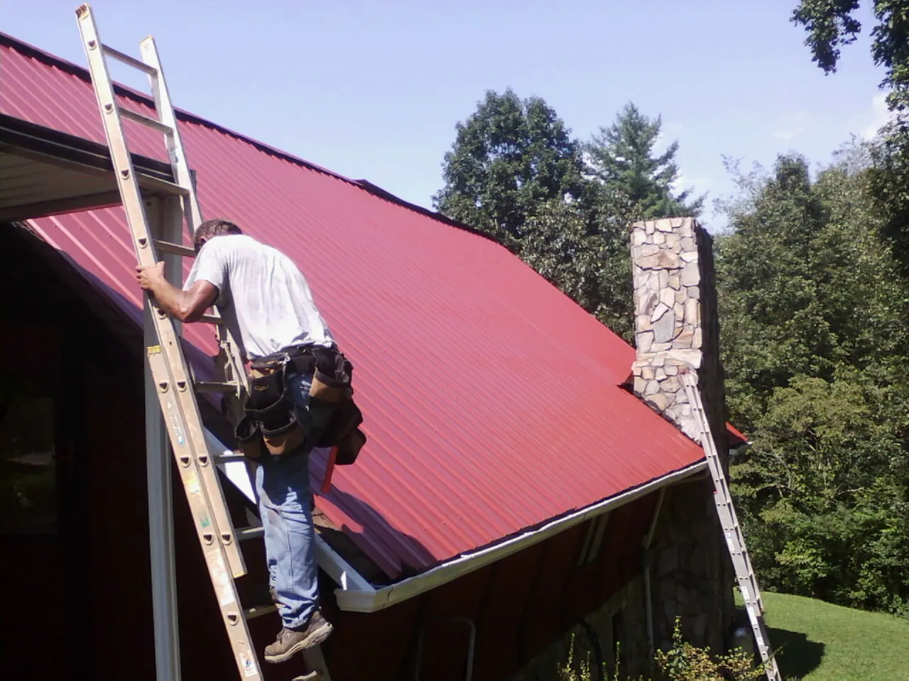 Roofer on a ladder beside a red metal roof and stone chimney — Soffit & Fascia Repair by Roofing Brothers in Bamberg